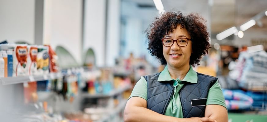 Store employee in green polo and vest smiles in convenience store with Worldwide Marketing logo. Product shelves in background.